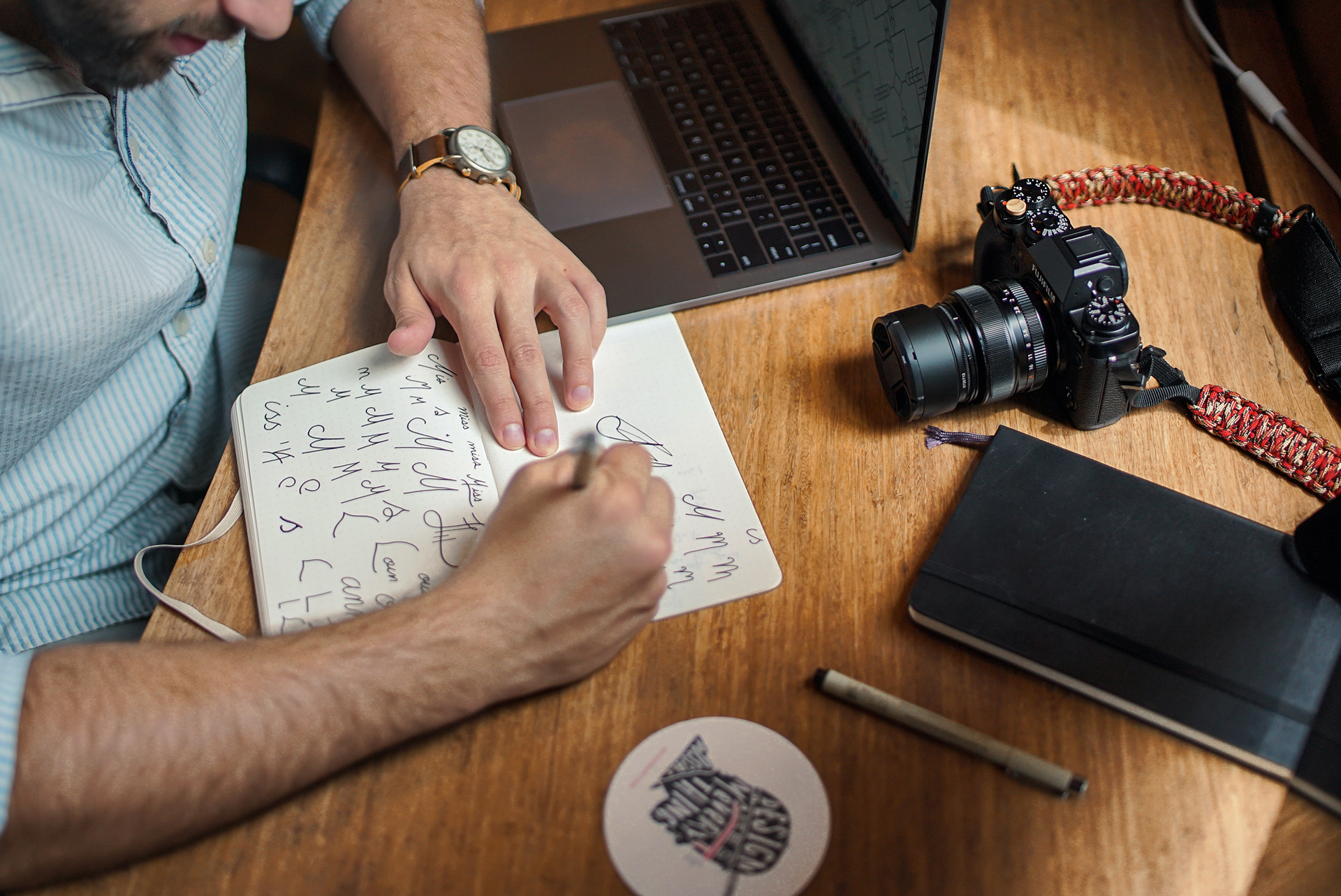 A person sitting at a wooden desk writing in a notebook with a silver pen, beside a laptop, a camera, and other photography equipment.