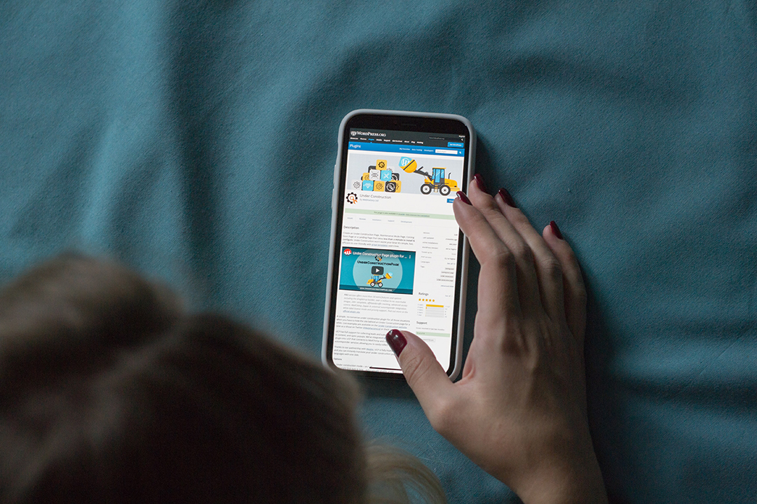 A person is lying on a blue blanket while holding a smartphone showcasing a web page with graphics and text.