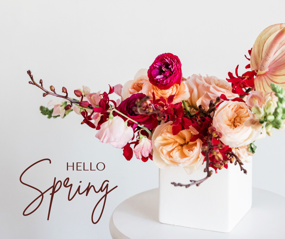 A vibrant arrangement of flowers in shades of red, pink, and peach presented in a white square vase on a white table, with the words "HELLO Spring" in cursive script to the bottom left corner of the image.