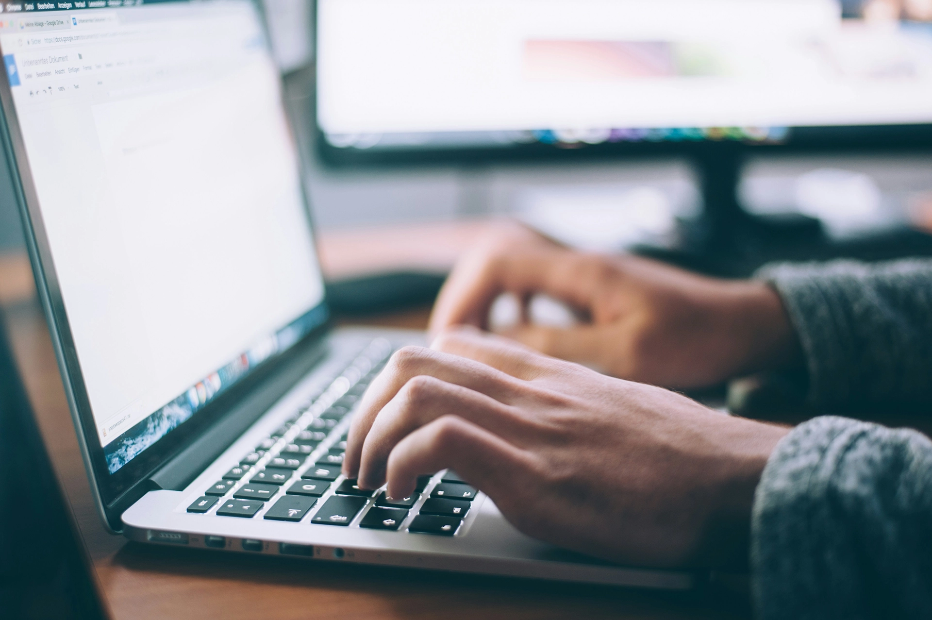 A close-up of a person's hands typing on a laptop keyboard with a blurred computer monitor in the background.